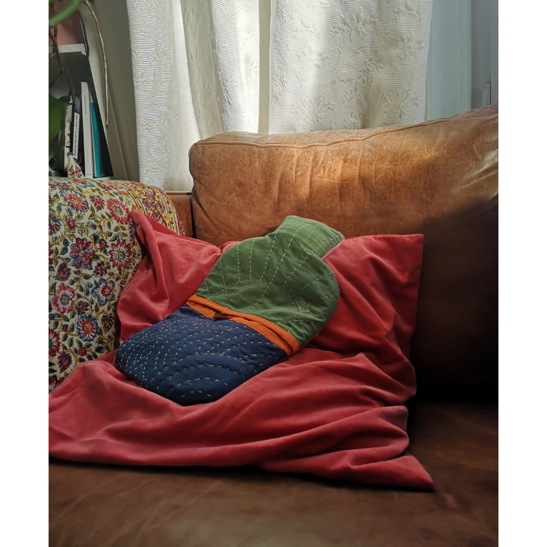 A hot water bottle lies on a pink cushion on brown leather chair. It is navy and green with orange bia binding across the front opening. It is hand quilted with an abstract sun on the top and semi circles on the bottom half. 