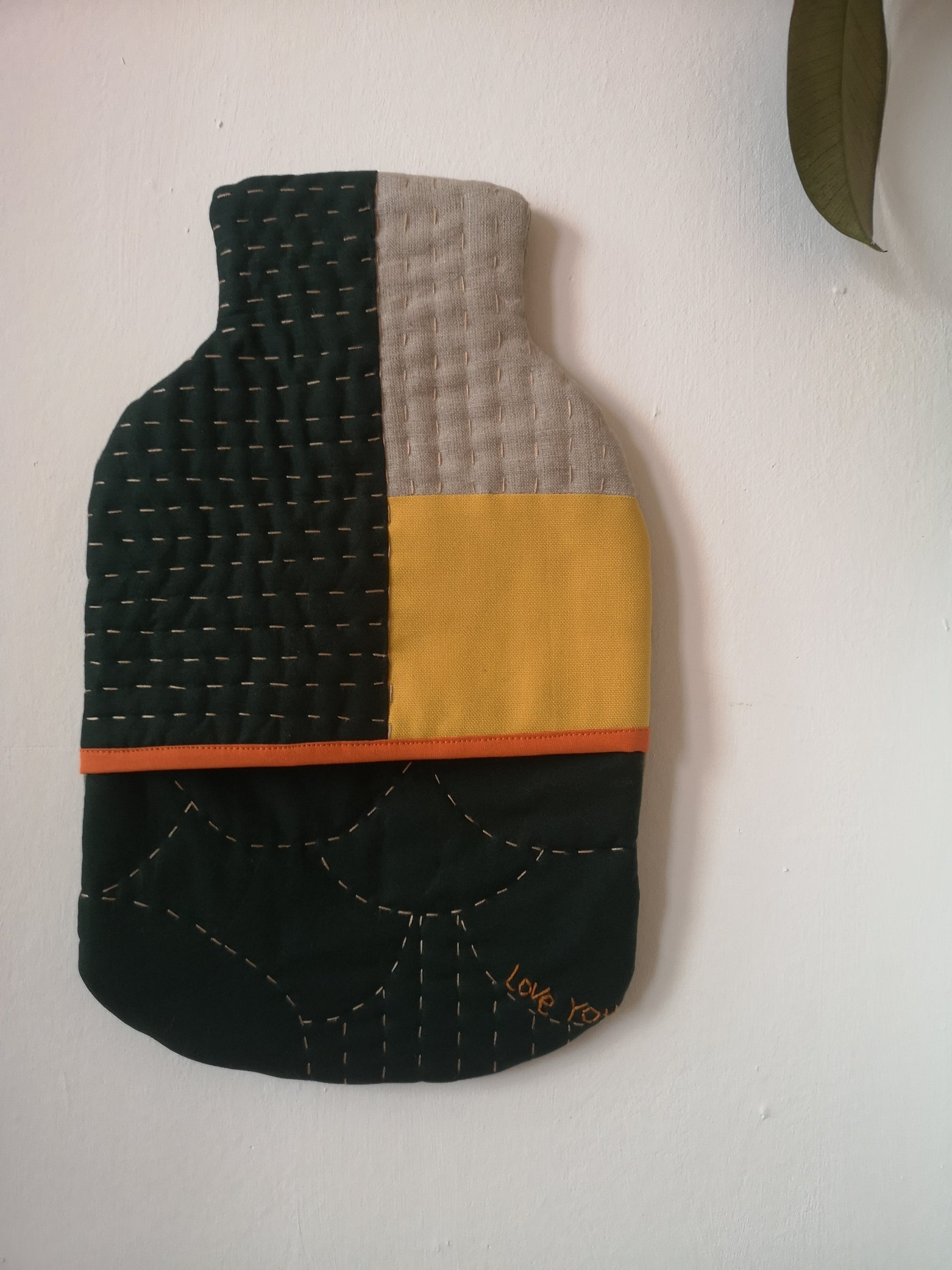 A hot water bottle cover on a white background, it is green, yellow and light grey with hand quilting and an orange binding across the front opening. There is a small embroidered message 'love you' on the bottom right corner. 