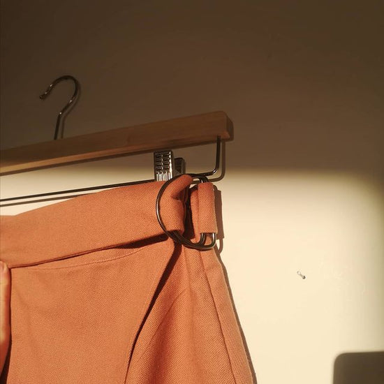 Close up of a pair of orange trousers on a wooden hanger in front of a white wall. 